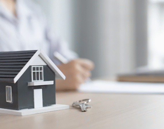 model home on desk with key to the right and background of person writing with pen and paper.