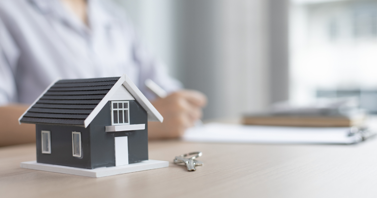 model home on desk with key to the right and background of person writing with pen and paper.