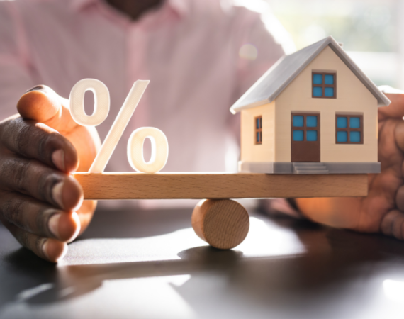 two hands open on desk balancing a seesaw with a % symbol on one side and minature house on the other